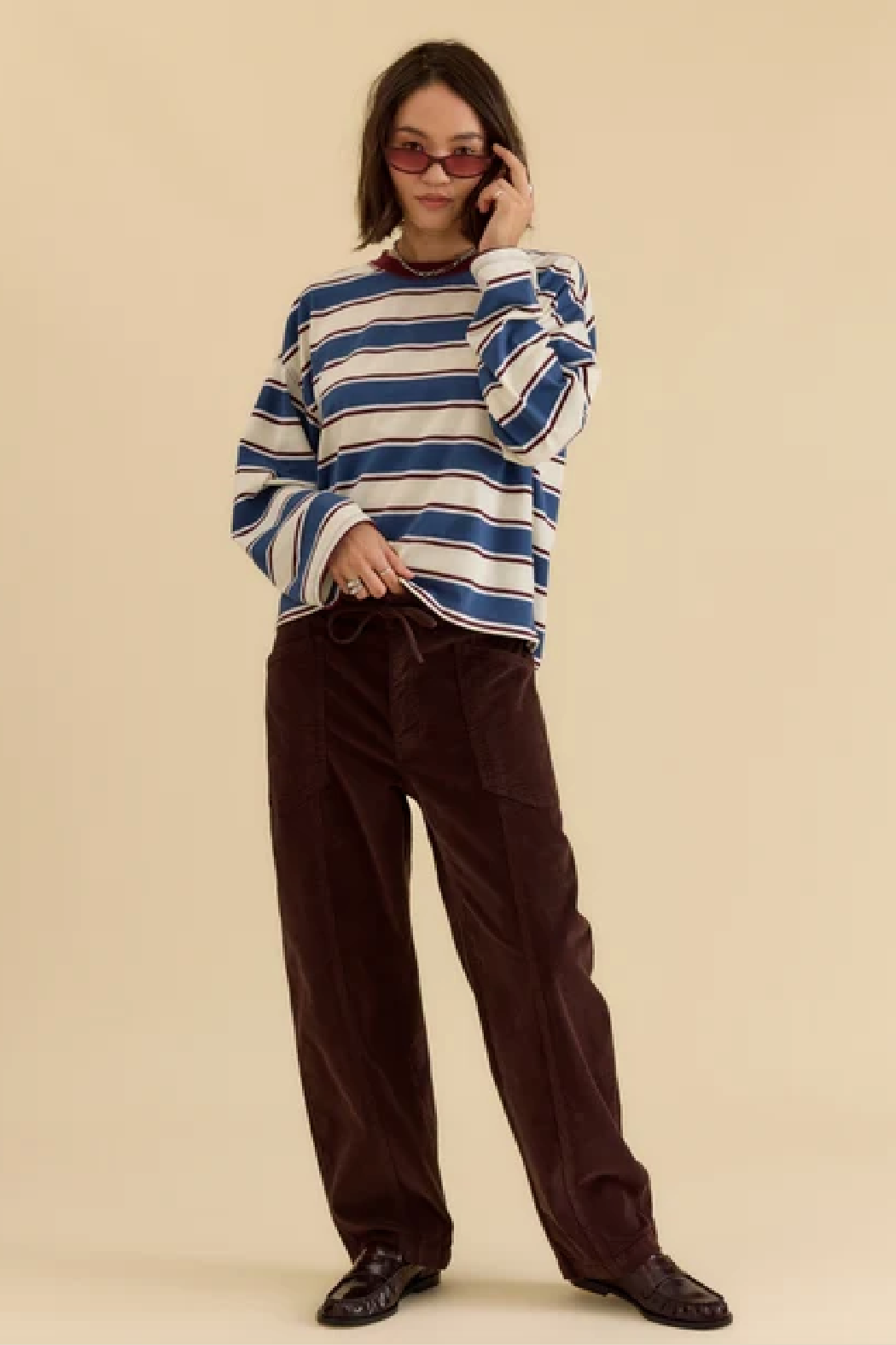 A model wearing a blue and white striped long-sleeve knit top, dark brown corduroy pants, and black leather loafers, standing against a solid beige studio background.