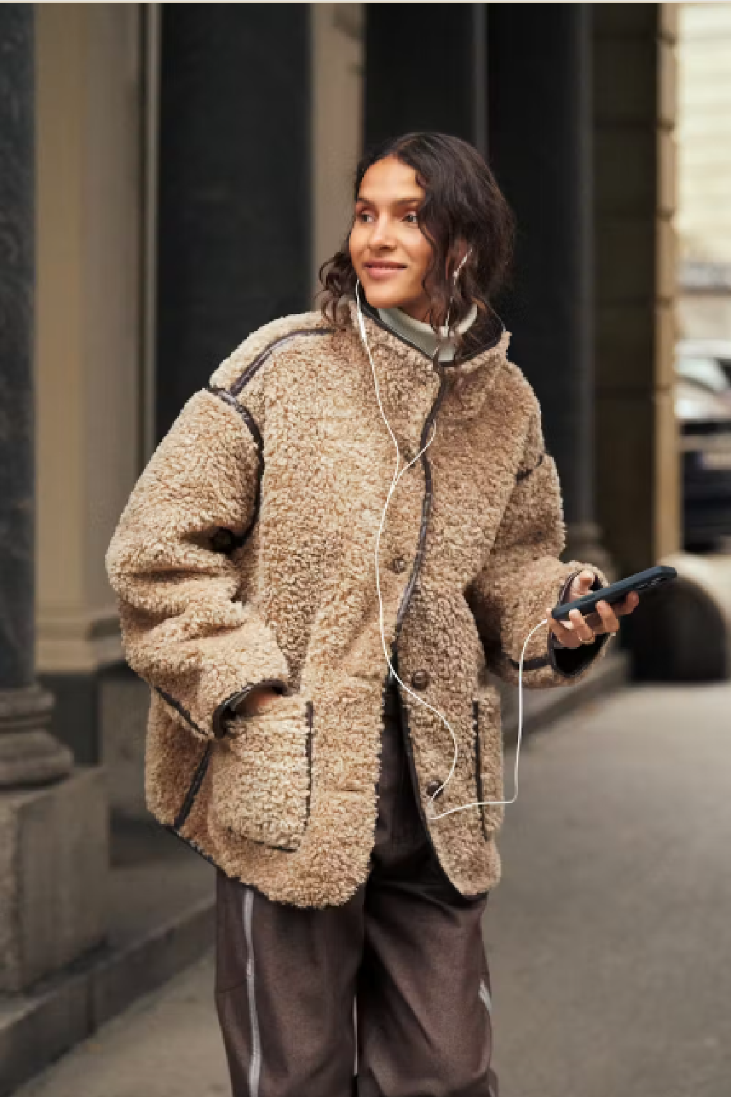 A woman walking on a city street wearing a beige sherpa fleece jacket with black piping, brown pants, and white wired earphones.