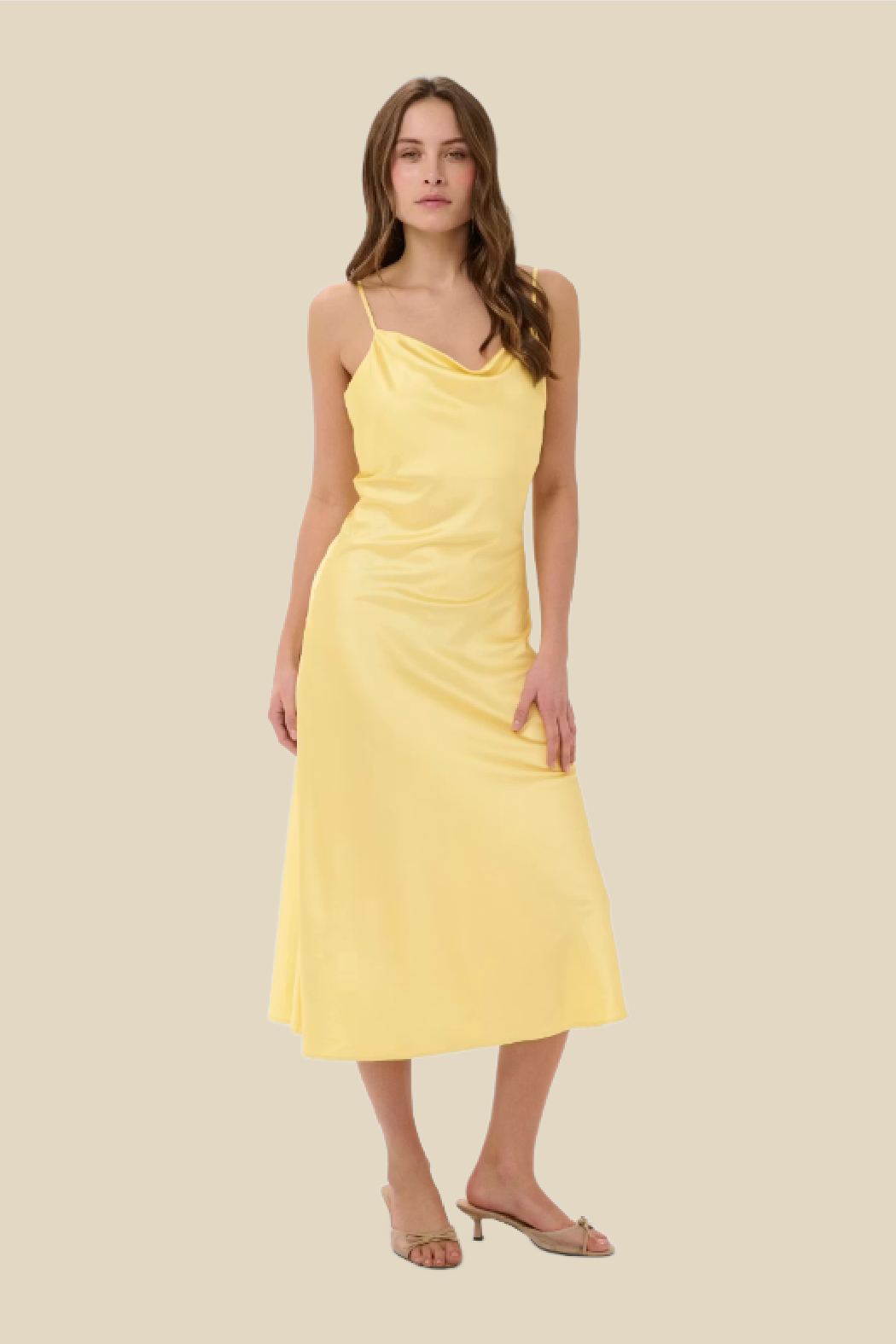 A woman modeling a pale yellow midi slip dress with thin straps, a draped neckline, and a gathered waist, paired with clear high-heel sandals against a solid beige background.
