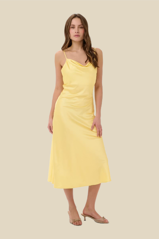 A woman modeling a pale yellow midi slip dress with thin straps, a draped neckline, and a gathered waist, paired with clear high-heel sandals against a solid beige background.