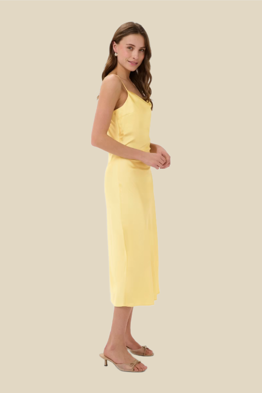 A woman modeling a pale yellow sleeveless midi dress with thin straps, standing against a solid beige background.