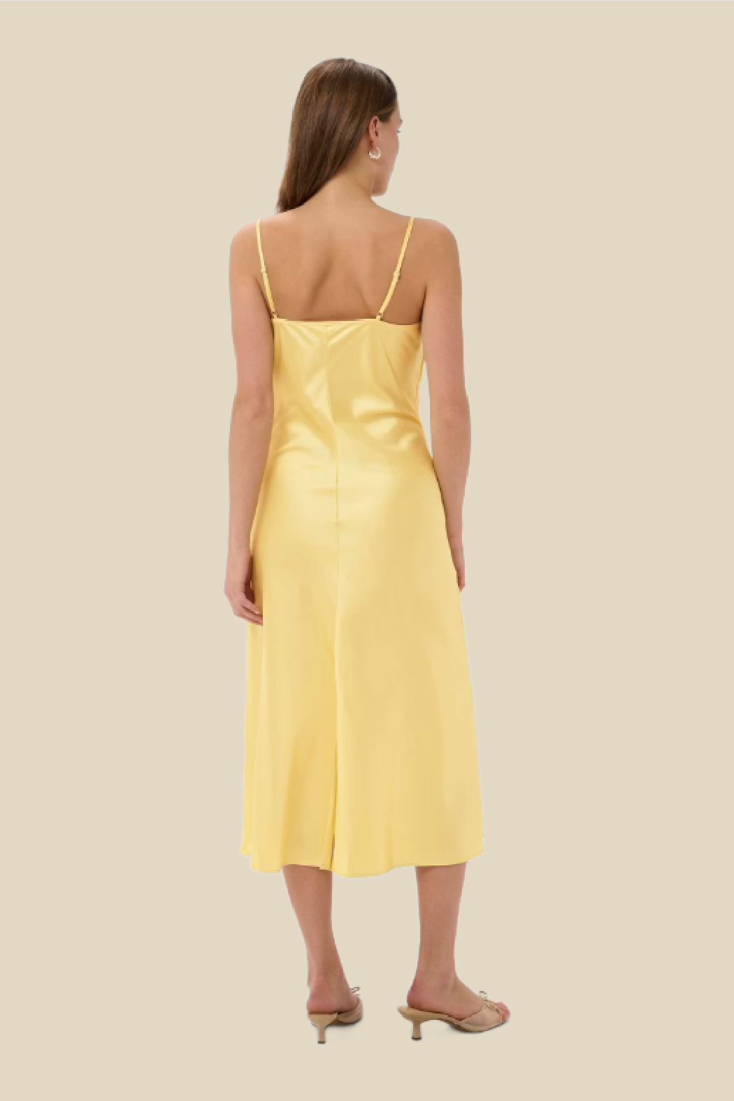 A woman viewed from the back wearing a pale yellow satin midi slip dress with thin spaghetti straps and nude heeled sandals, set against a solid light beige background.