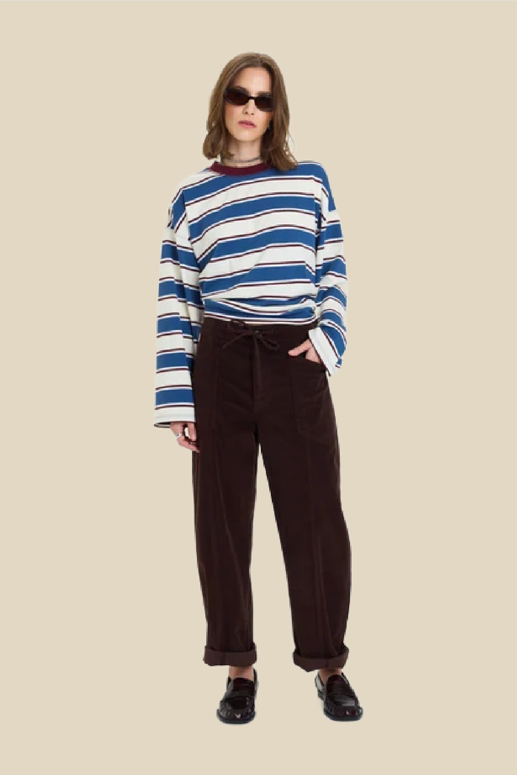 A model wearing a blue, white, and navy striped long-sleeve knit top, dark brown corduroy pants, and black loafers, standing against a solid beige background.