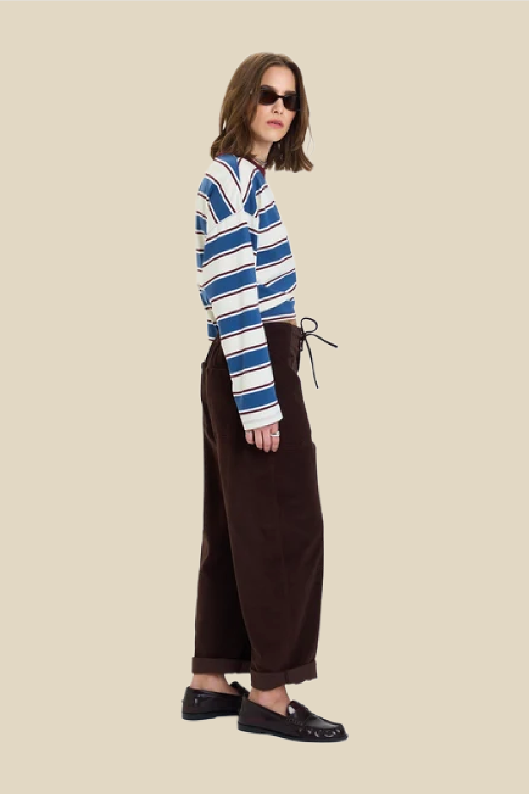 A model wearing a blue and white striped long-sleeve top, dark brown wide-leg corduroy pants, and black loafers, posed against a solid beige background.