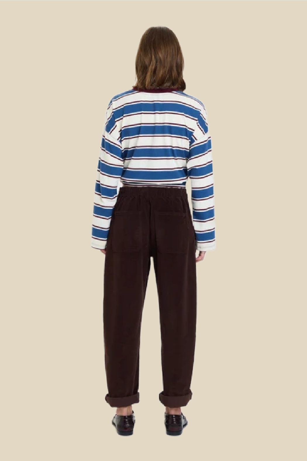 A model viewed from the back wearing a blue, white, and navy striped long-sleeve crop top and dark brown corduroy trousers, set against a solid beige background.