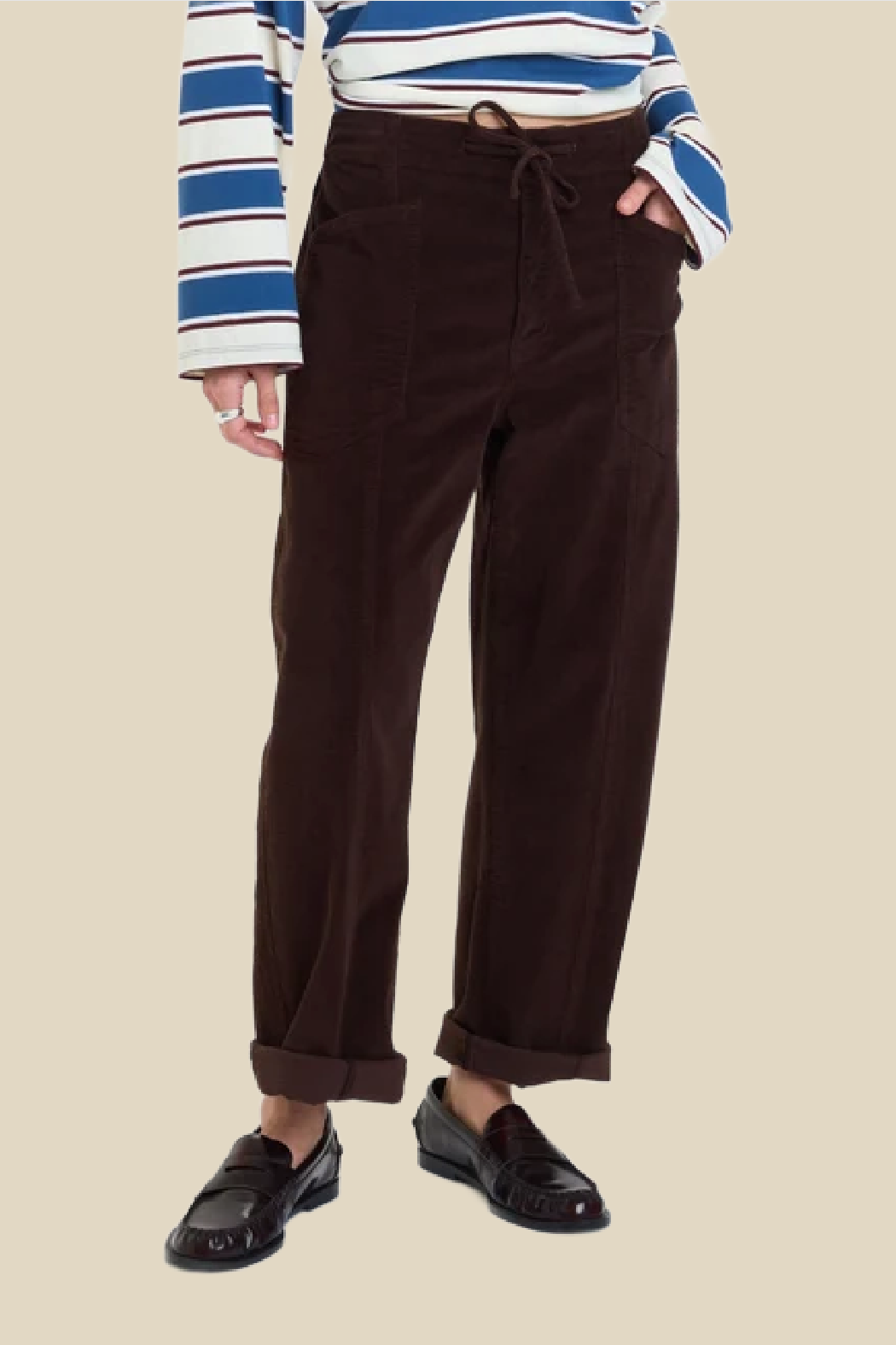 A person wearing dark brown corduroy drawstring pants, a blue and white striped long-sleeve top, and black loafers, shown from the chest down against a plain beige background.