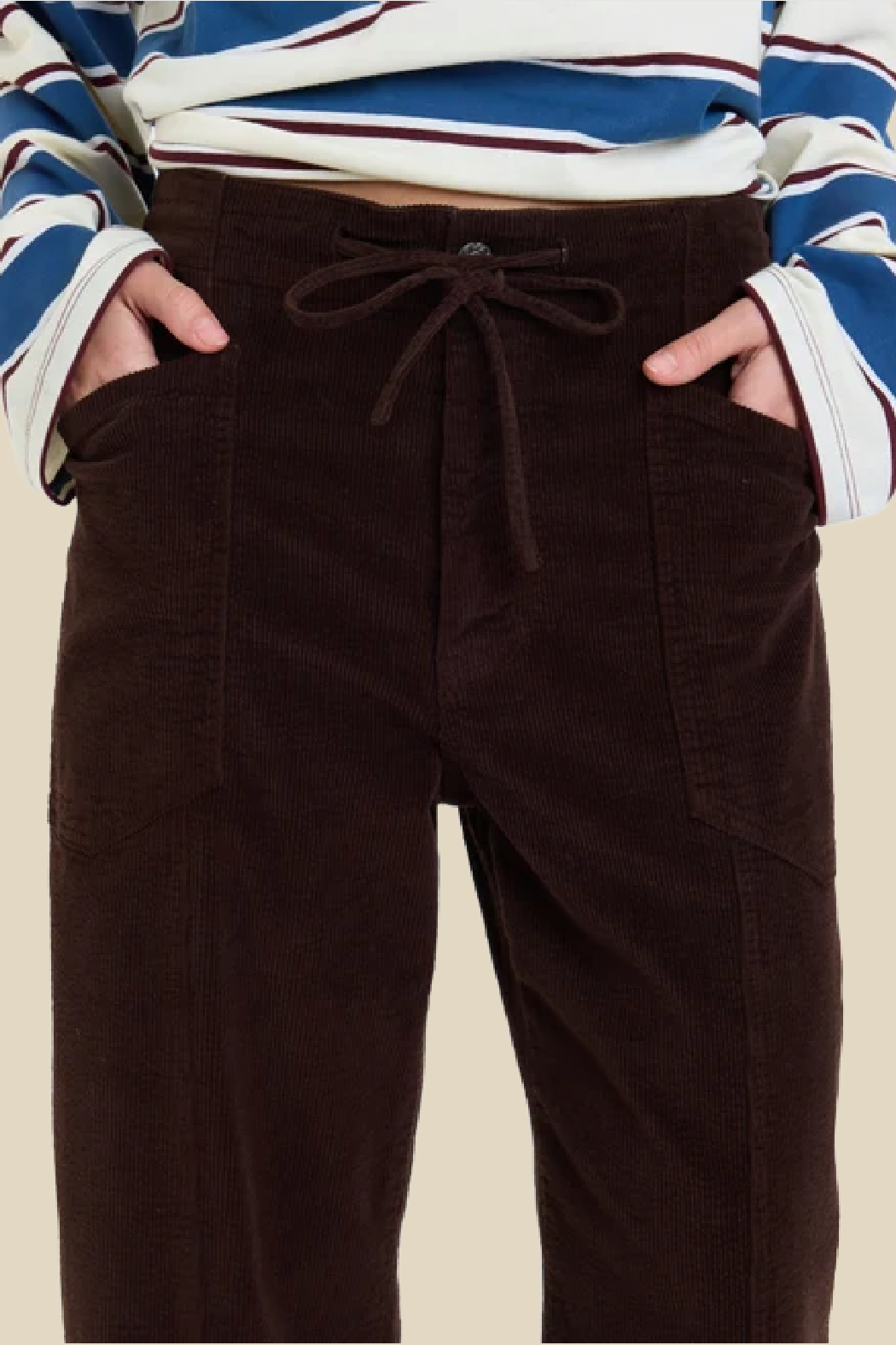Dark brown corduroy pants with a drawstring waist and side pockets, worn by a model with a blue, white, and maroon striped long-sleeve top against a light beige background.