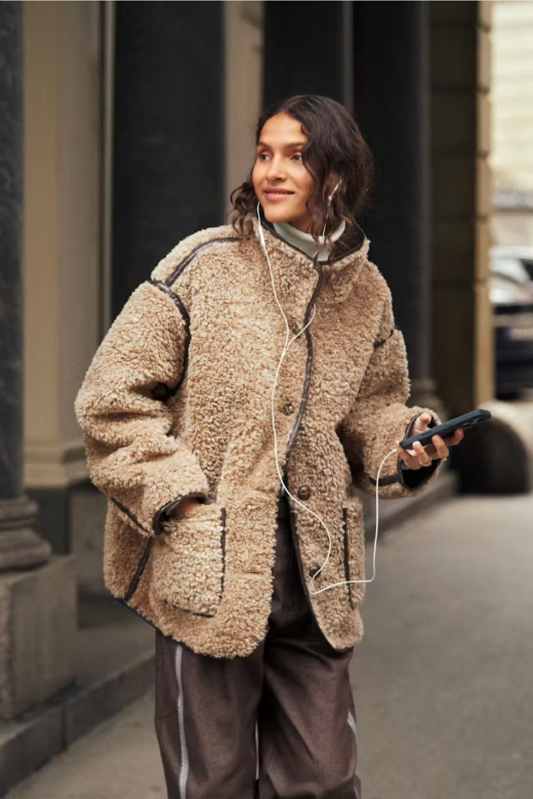 A woman wearing a beige sherpa fleece jacket with black contrast stitching, brown pants, and white wired earphones, walking on a city street while holding a smartphone.