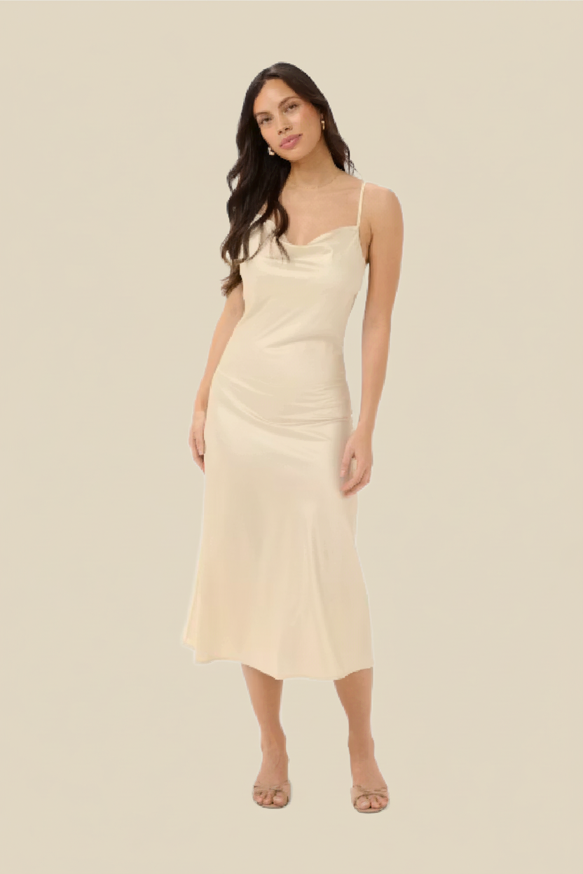 A woman modeling a light cream satin slip dress with spaghetti straps and a draped neckline, set against a solid beige background.
