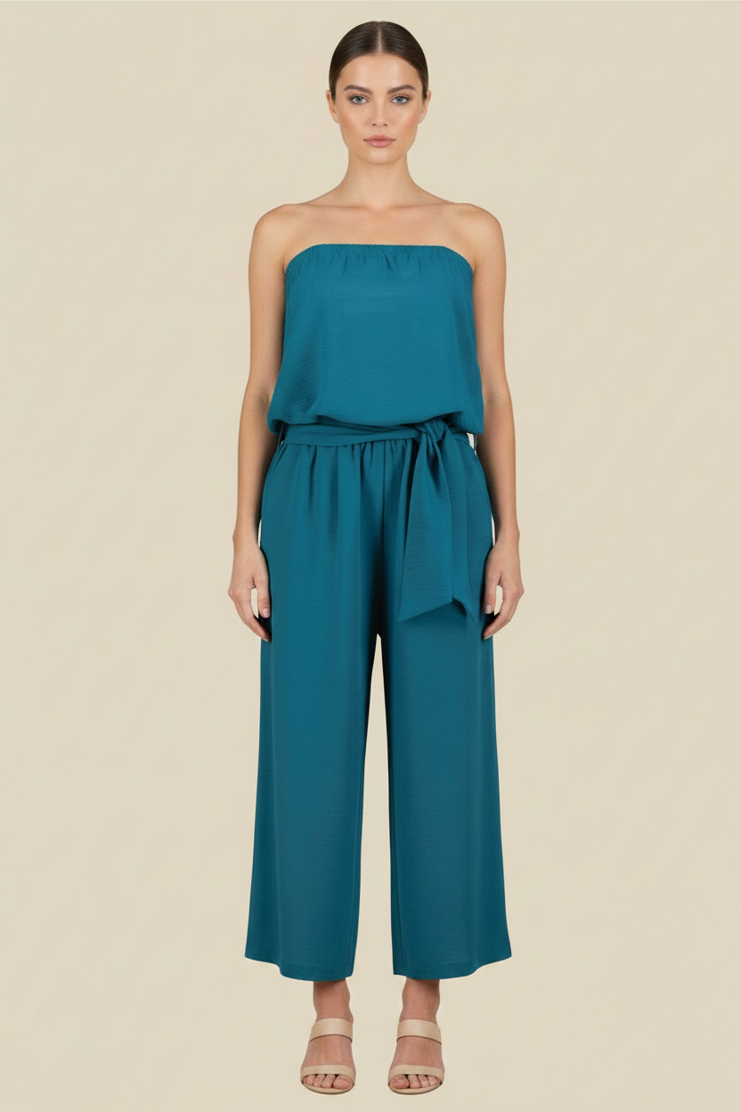 A woman modeling a teal strapless jumpsuit with a gathered top and wide-leg pants, standing against a solid beige background.