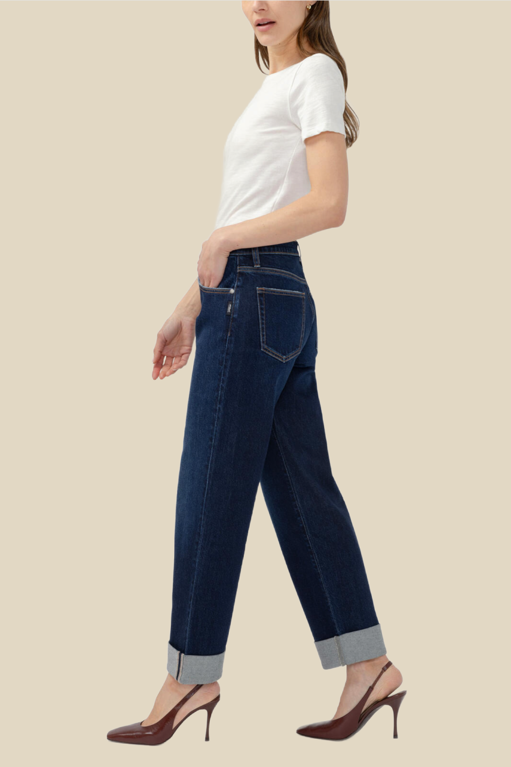 A woman shown in side profile wearing dark blue straight-leg jeans with rolled cuffs, a white short-sleeve top, and brown pointed-toe heels against a plain beige studio background.