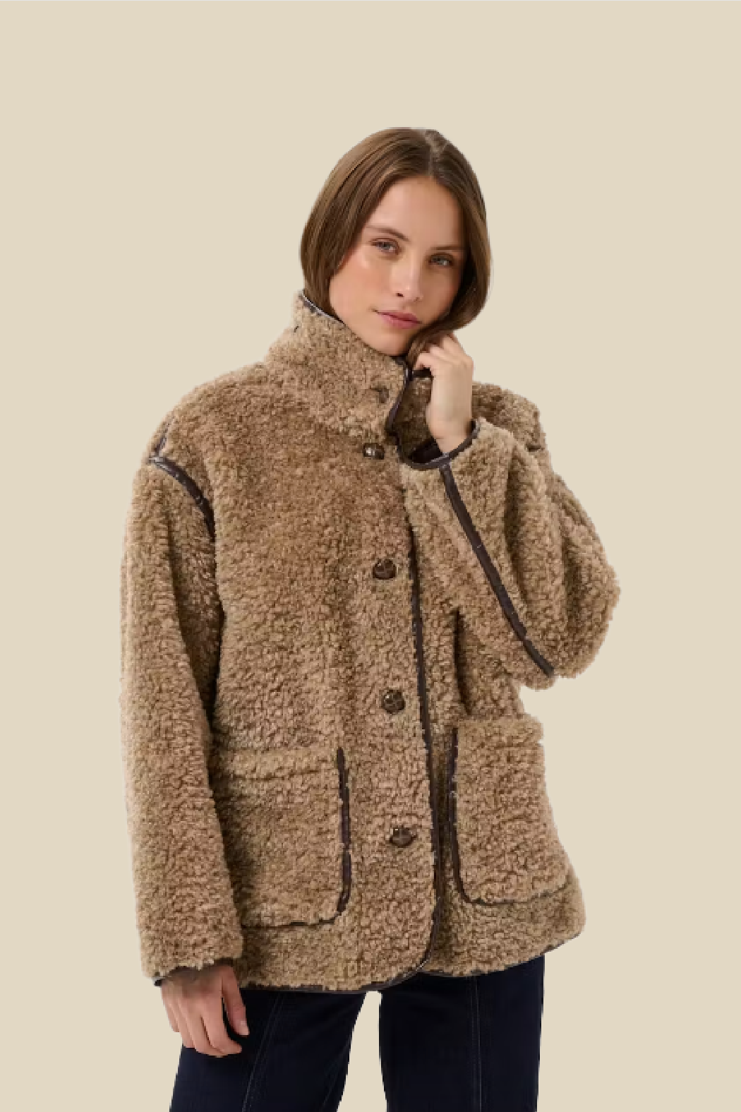 A woman wearing a brown curly sherpa fleece jacket with dark contrast piping and front buttons, paired with dark blue jeans, against a solid beige background.