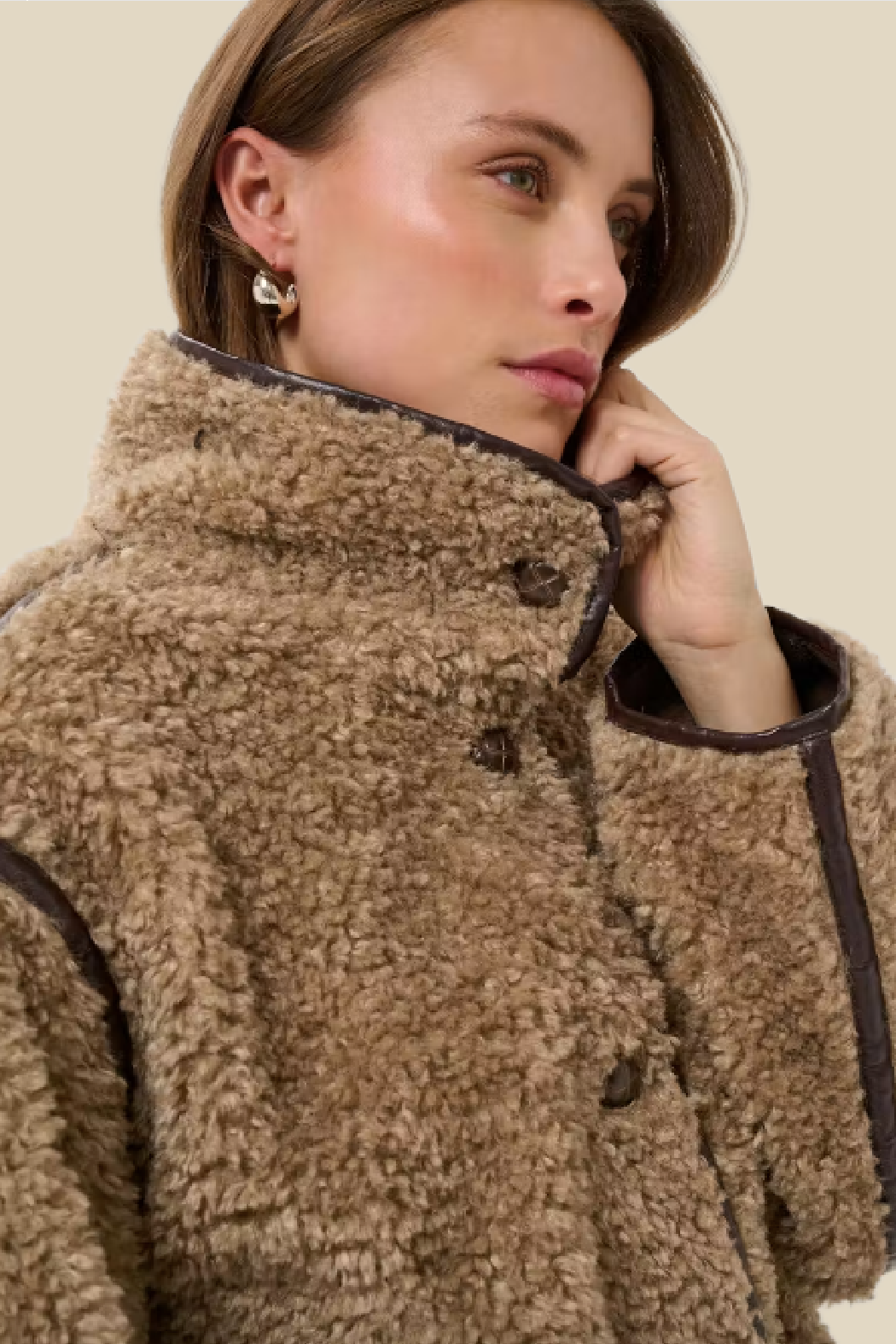A close-up of a woman wearing a brown sherpa fleece jacket with dark brown trim and buttons, set against a solid light beige background.