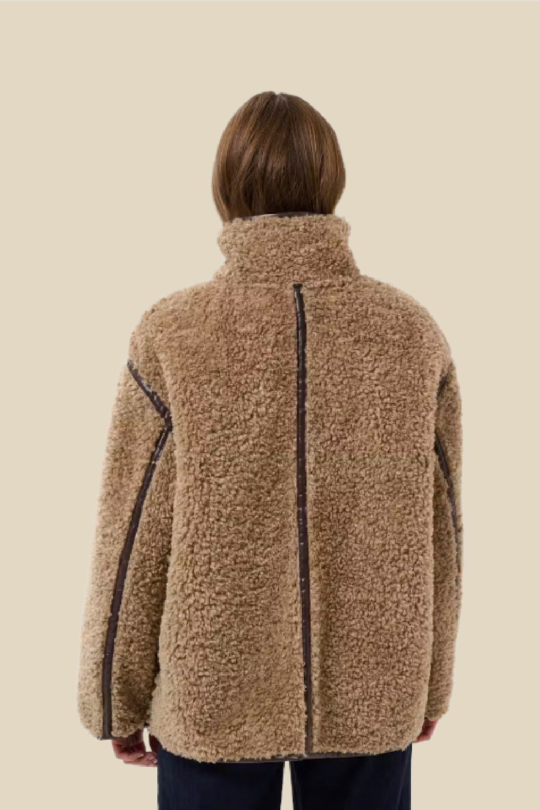 A person shown from the back wearing a beige sherpa fleece jacket with dark contrast stitching, set against a solid light beige background.