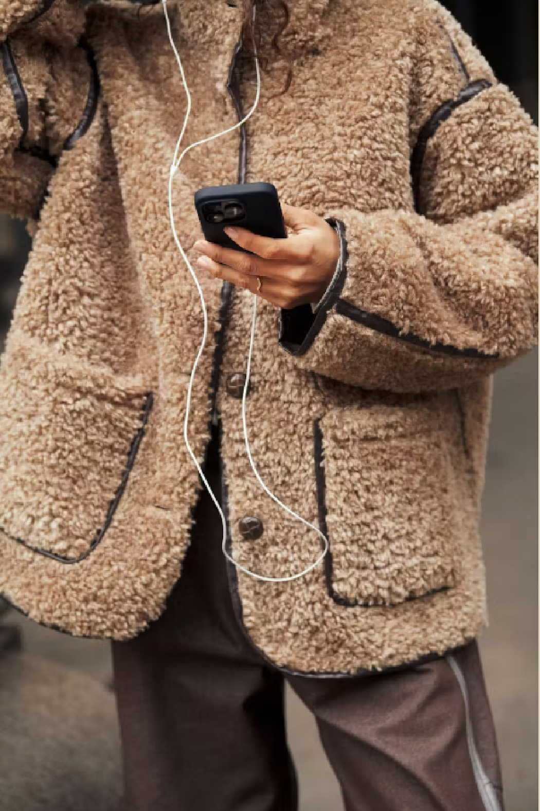 A person wearing a beige sherpa fleece jacket with black contrast trim, holding a black smartphone with white wired earphones. The jacket has large front patch pockets and a button-front closure.