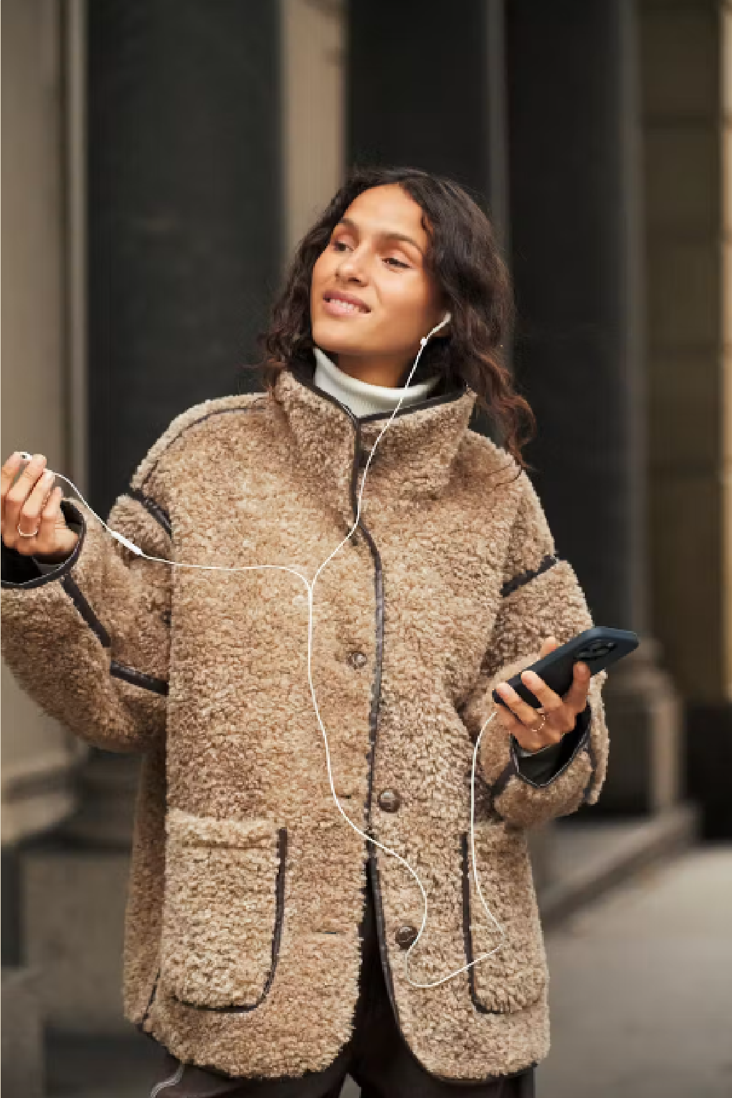 A woman wearing a beige sherpa fleece jacket with black trim, standing outdoors near columns and holding white wired earphones and a smartphone.