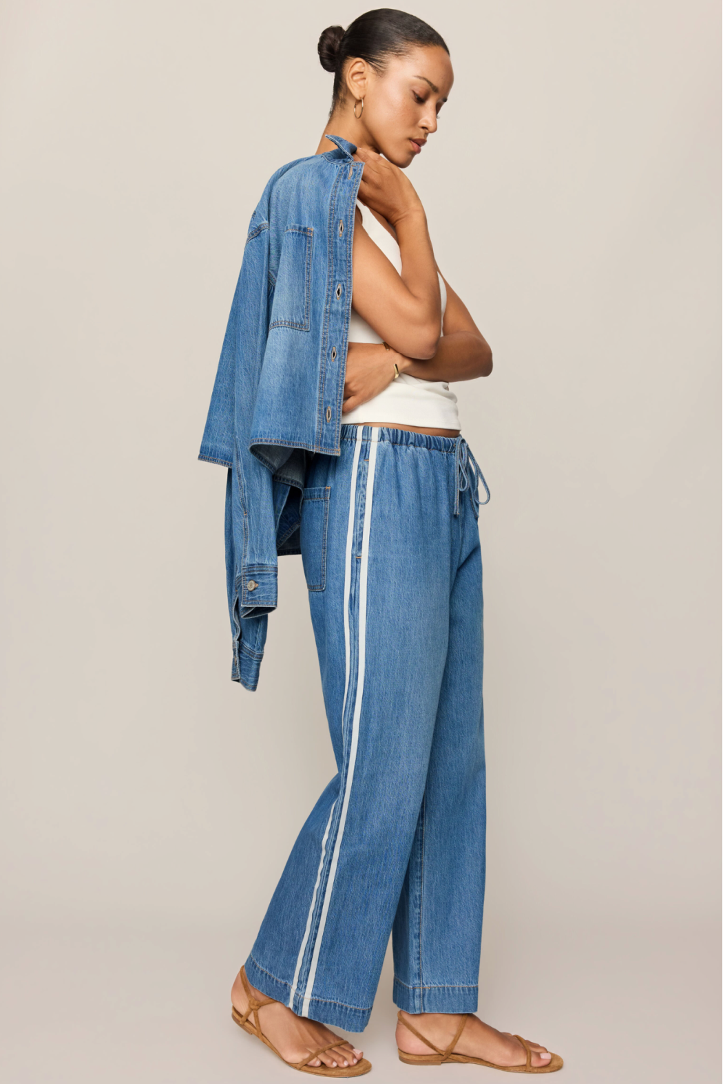 A woman modeling a pair of wide-leg blue denim pants with white side stripes, styled with an oversized blue denim shirt draped off her shoulders and a white tank top, against a solid light beige background.