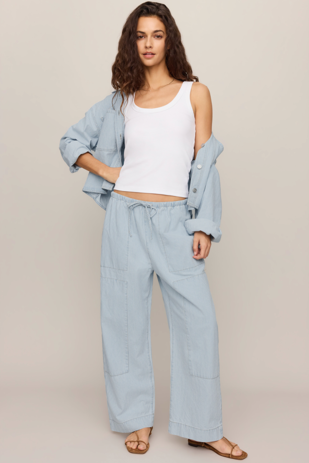 A woman modeling a light blue wide-leg linen pants set, featuring a matching drawstring waist and large patch pockets, styled with a white ribbed tank top and an open light blue button-up shirt.